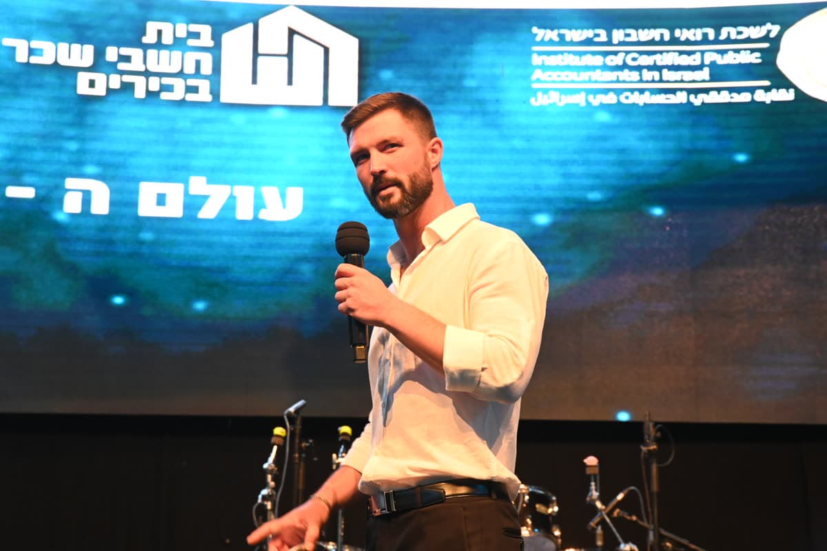מקסים גומברג עם מיקרופון בכנס חשבי שכר בכירים, מול מסך ״עולם ה-AI״