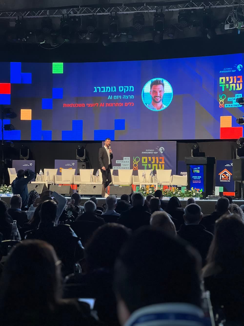 מקסים גומברג על במת ועידת ״בונים עתיד״, שמו ותמונתו על המסך, האולם מלא